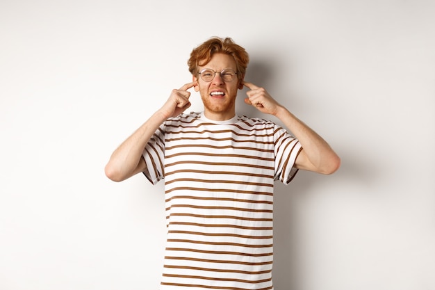 Annoyed redhead guy in glasses shutting ears and complaining loud noise, angry at noisy neighbours, standing over white background.