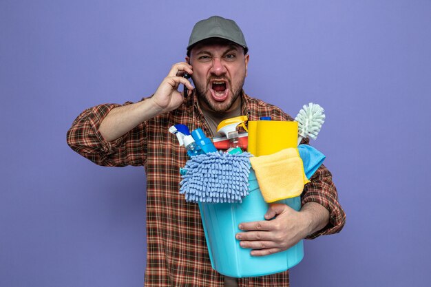 Раздраженный славянский уборщик держит оборудование для уборки и кричит на кого-то по телефону