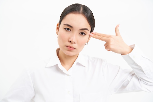 Free photo annoyed and tired asian businesswoman shows pistol gesture on head looks bothered and tensed stands