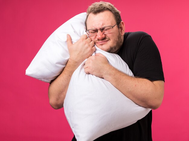 Annoyed with closed eyes middle-aged ill male hugged pillow isolated on pink wall