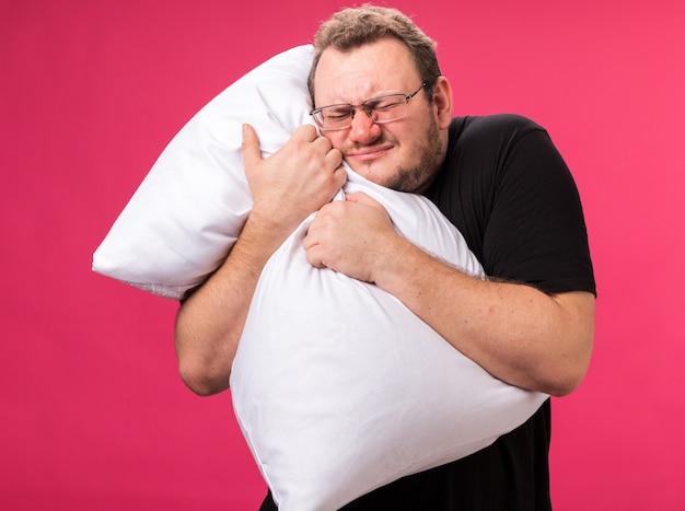 Раздраженный с закрытыми глазами больной мужчина средних лет обнял подушку, изолированную на розовой стене