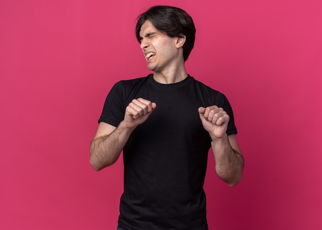 Annoyed with closed eyes young handsome guy wearing black t-shirt holding fists isolated on pink wall