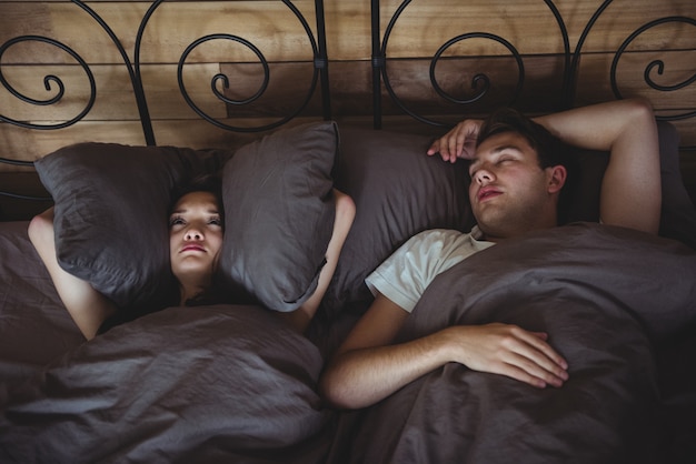 Annoyed woman covering her ears with pillows to block out snoring in bedroom