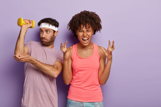Annoyed woman gestures angrily, can't continue training and hard working guy works on having muscles, dressed in sportwear