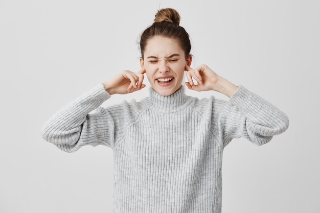 Annoyed woman plugging ears with fingers and screwing up her eyes in displeasure. Brunette female covering ears complaining on loud music from outside. Human feelings, reactions