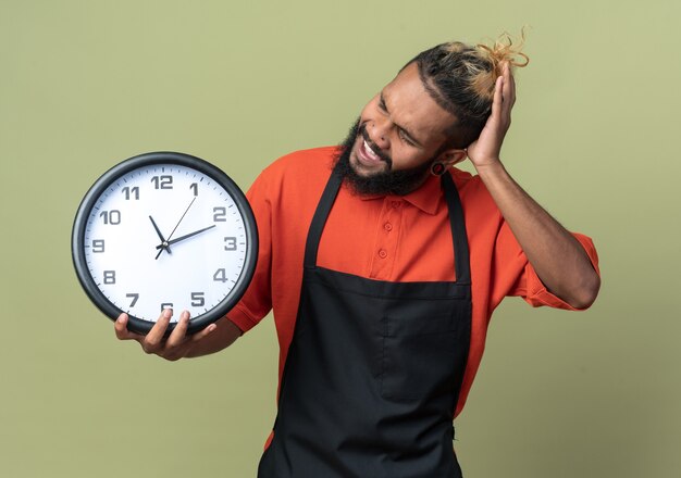 Annoyed young afro-american barber wearing uniform holding and looking at clock keeping hand on head isolated on olive green wall