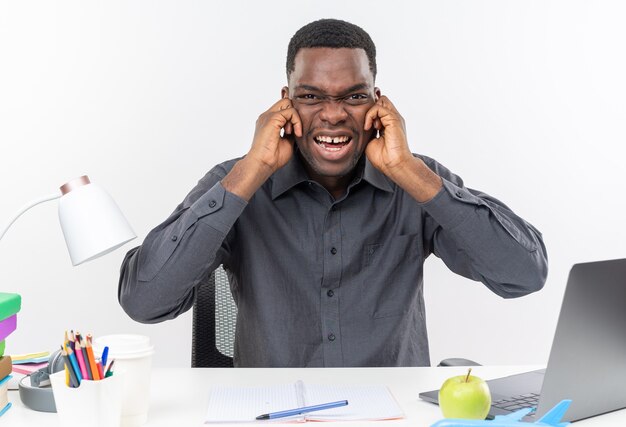 Annoyed young afro-american student sitting at desk with school tools closing his ears with fingers isolated on white wall