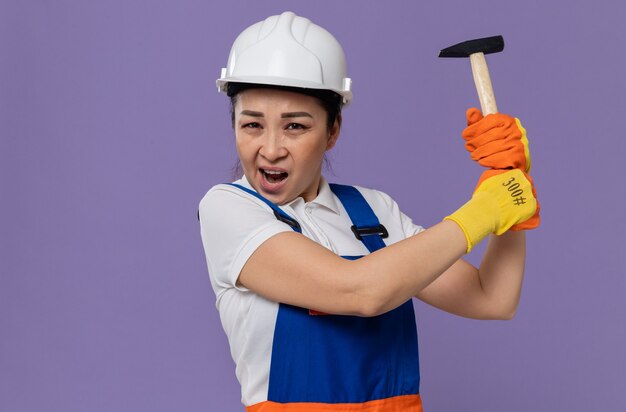 Раздраженная молодая азиатская женщина-строитель с белым защитным шлемом и перчатками, держащая молоток