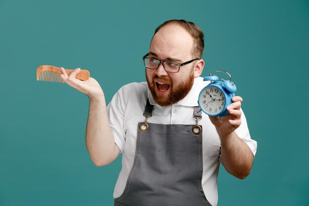 Annoyed young barber wearing uniform and glasses looking at side showing alarm clock holding comb in another hand isolated on blue background