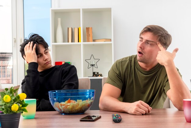 annoyed young blonde and brunette handsome guys sit at table with bowl of chips phone and tv remote putting hand on head looking at each other