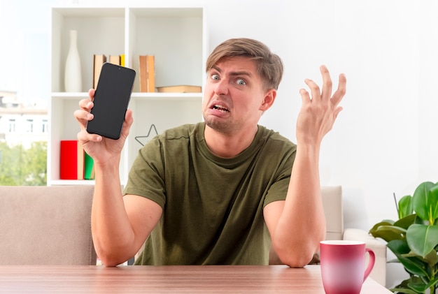 Annoyed young blonde handsome man sits at table raising hand and holding phone inside the living room