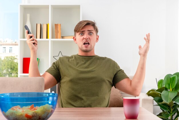 Annoyed young blonde handsome man sits at table with bowl of chips and cup raising hands up holding tv remote inside the living room