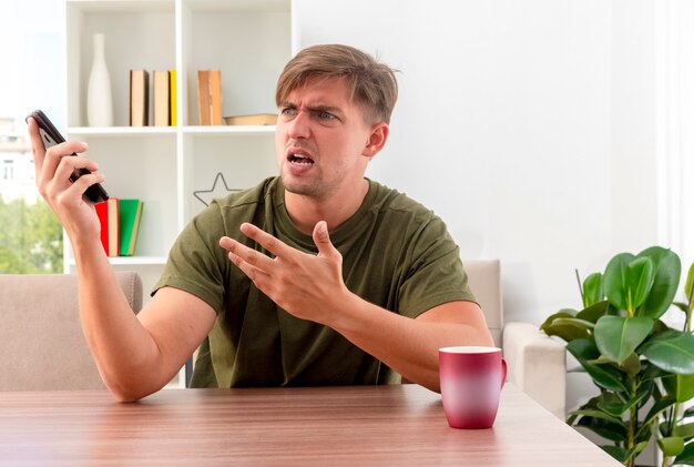 Annoyed young blonde handsome man sits at table with cup holding phone looking and pointing at side with hand inside living room