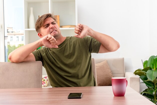 Annoyed young blonde handsome man sits at table with cup and phone keeping fists looking at side inside the living room