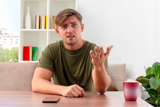 Annoyed young blonde handsome man sits at table with cup and phone looking and pointing  with hand