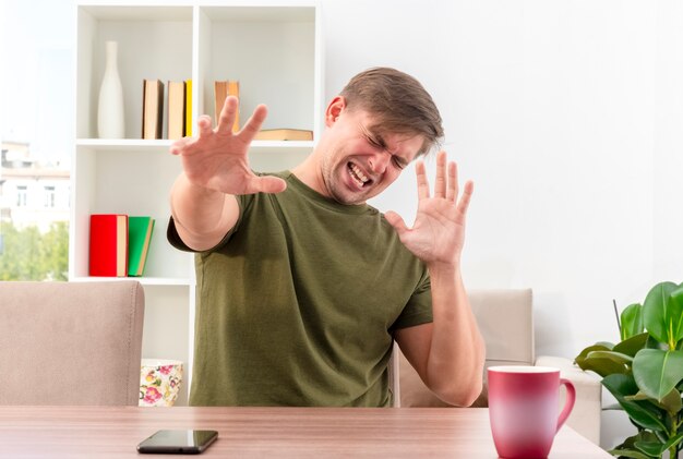 Annoyed young blonde handsome man sits at table with cup and phone raising hands inside living room