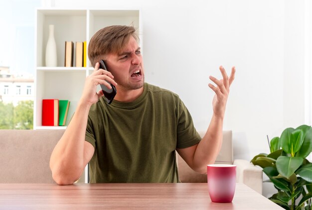 Раздраженный молодой блондин красавец сидит за столом с чашкой, кричит на кого-то по телефону с поднятой рукой, глядя в сторону