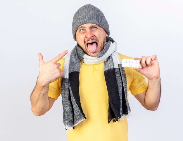 Annoyed young blonde ill man wearing winter hat and scarf holds pack of medical pills and points at side isolated on white wall