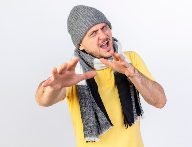 冬の帽子と白い壁で隔離の正面を見て手を伸ばしてスカーフを身に着けているイライラする若い金髪の病気の男
