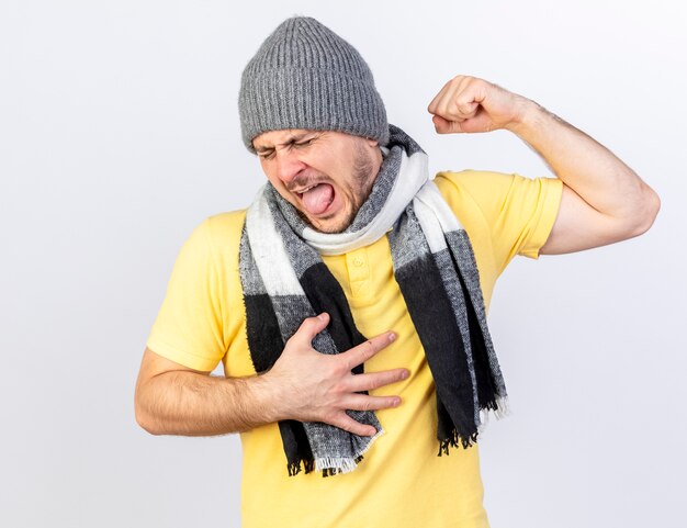 Annoyed young blonde ill man wearing winter hat and scarf tenses biceps puts hand on chest isolated on white wall