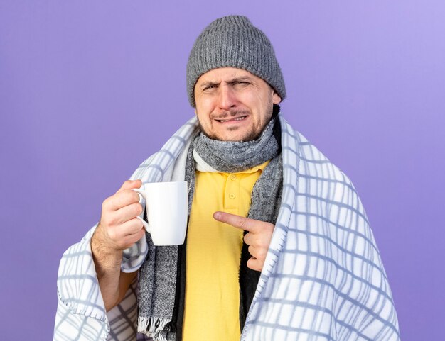 Annoyed young blonde ill slavic man wearing winter hat and scarf wrapped in plaid holds and points at cup isolated on purple wall with copy space