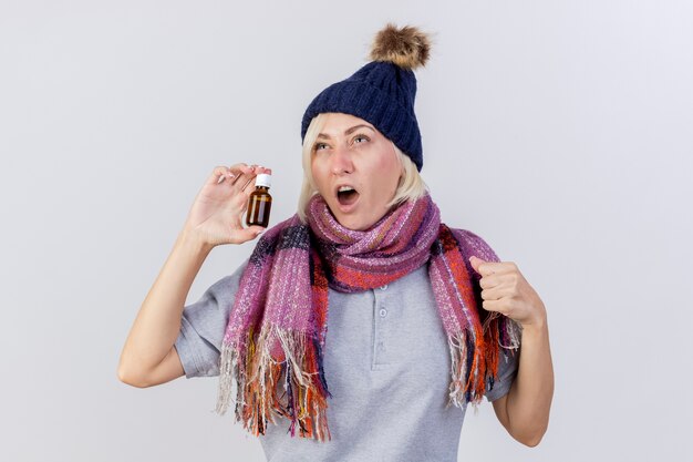 Раздраженная молодая блондинка больна славянская женщина в зимней шапке и шарфе держит лекарство в стеклянной бутылке и держит кулак, глядя вверх изолированно на белой стене с копией пространства