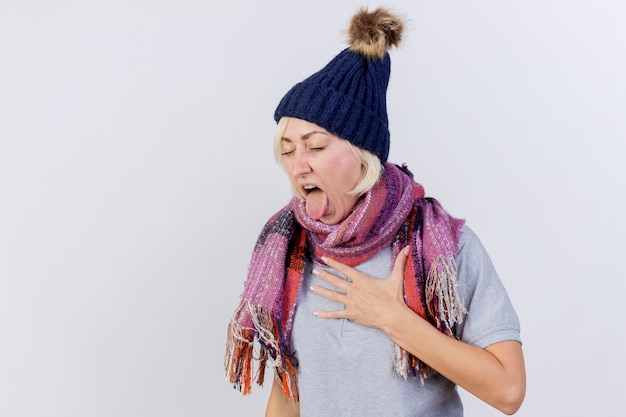 La giovane donna slava malata bionda infastidita che indossa il cappello e la sciarpa di inverno stuks fuori la lingua mette la mano sul petto isolata sulla parete bianca con lo spazio della copia