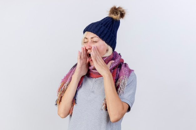 Free photo annoyed young blonde ill woman wearing winter hat and scarf stucks out tongue holding hands close to face isolated on white wall