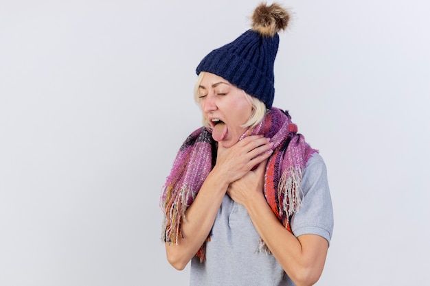 La giovane donna bionda malata infastidita che indossa il cappello e la sciarpa di inverno sporge la lingua e mette le mani sul collo isolato sulla parete bianca