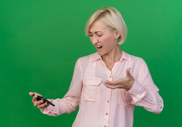 Annoyed young blonde slavic woman holding pointing at and looking at mobile phone isolated on green background