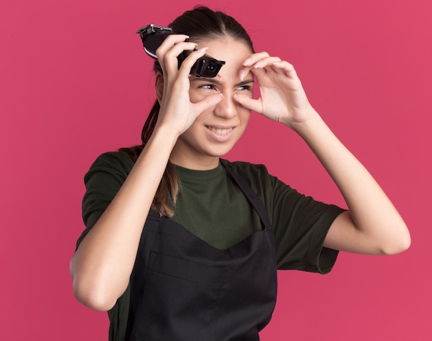 Annoyed young brunette barber girl in uniform holds hair clippers and looks at side through fingers on pink