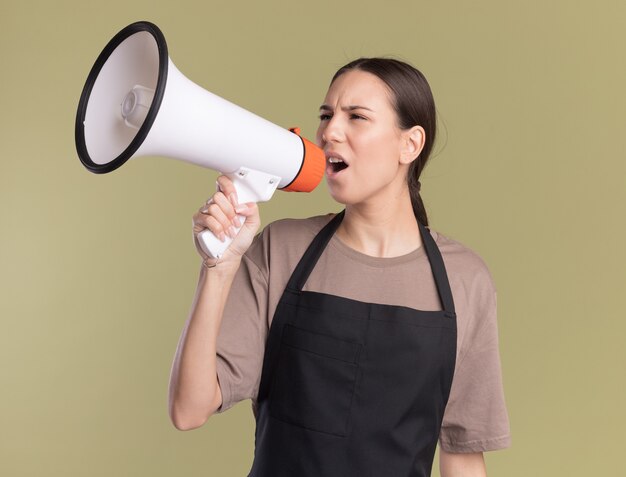 Annoyed young brunette barber girl in uniform holds and speaks into loud speaker on olive green