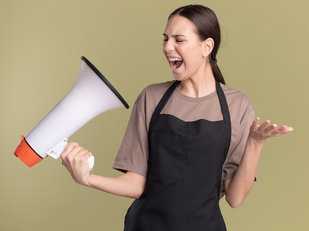 Annoyed young brunette barber girl in uniform keeps hand open holding and looking at loud speaker on olive green