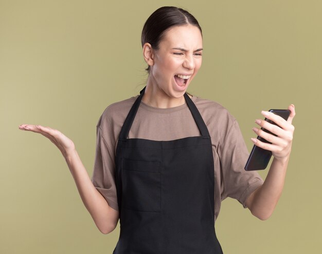 Annoyed young brunette barber girl in uniform keeps hand open holding and looking at phone on olive green