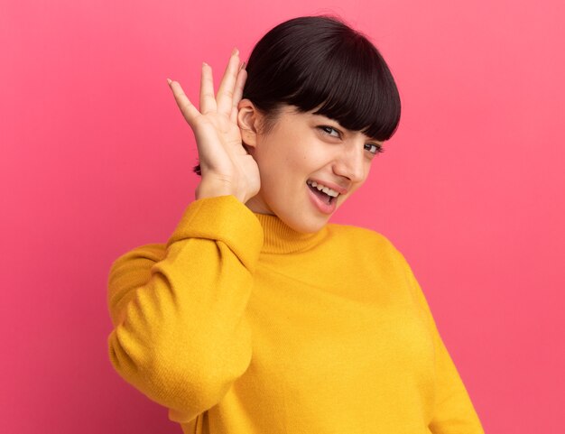 Annoyed young brunette caucasian girl holds hand behind ear looking at camera on pink