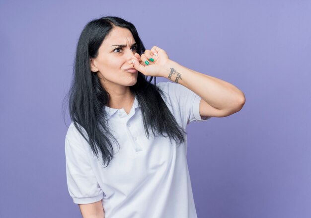 Annoyed young brunette caucasian girl holds nose looking at side isolated on purple wall