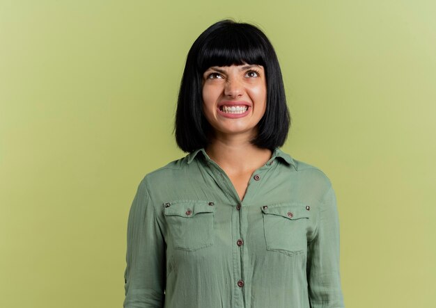 Annoyed young brunette caucasian girl looks up isolated on olive green background with copy space