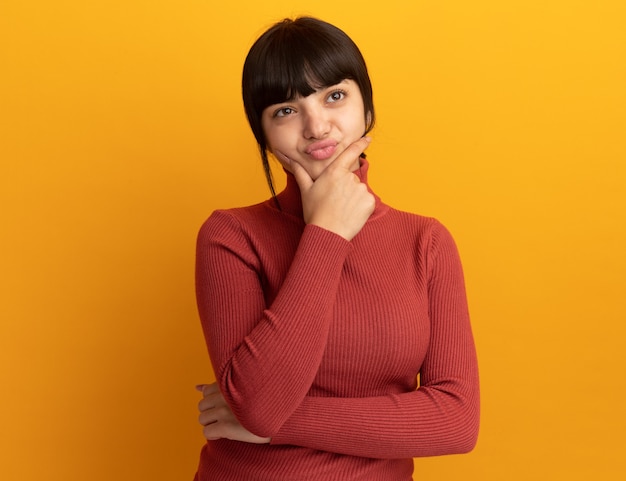 La giovane ragazza caucasica castana infastidita mette la mano sul mento e guarda al lato isolato sulla parete arancione con lo spazio della copia