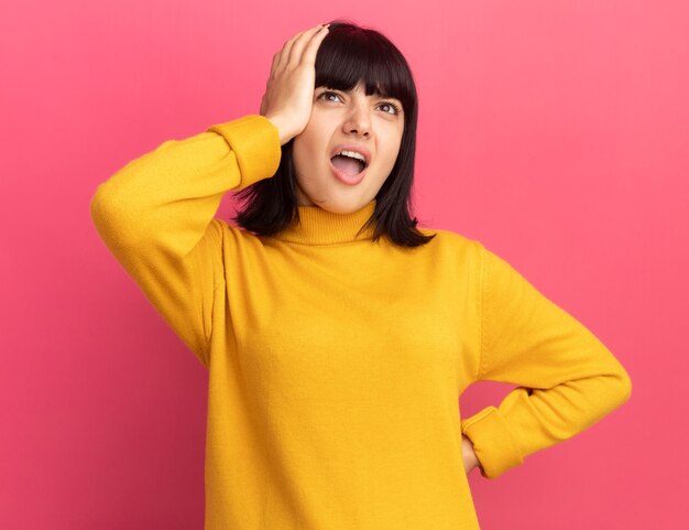 Annoyed young brunette caucasian girl puts hand on head and looks up on pink