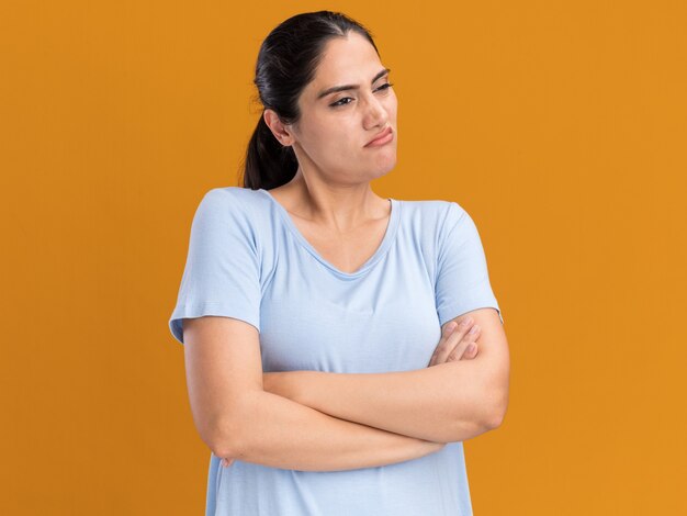 オレンジ色の側を見て腕を組んで立っているイライラする若いブルネットの白人の女の子