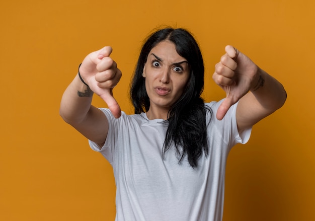 Annoyed young brunette caucasian girl thumbs down isolated on orange wall