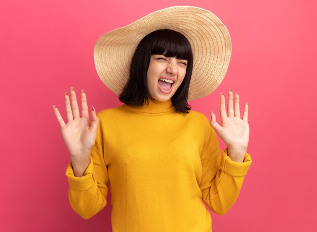Annoyed young brunette caucasian girl wearing beach hat stands with raised hands on pink 