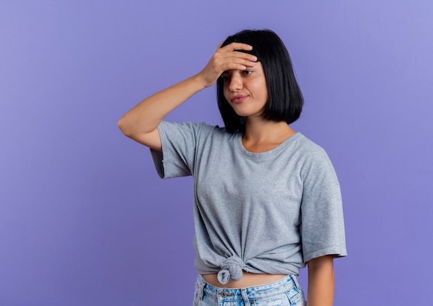 Annoyed young brunette caucasian woman puts hand on forehead looking at side isolated on purple background with copy space
