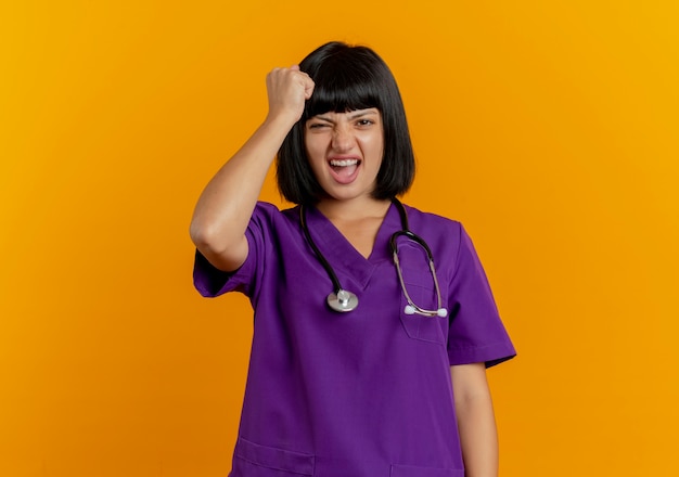 Annoyed young brunette female doctor in uniform with stethoscope puts fist on head isolated on orange background with copy space