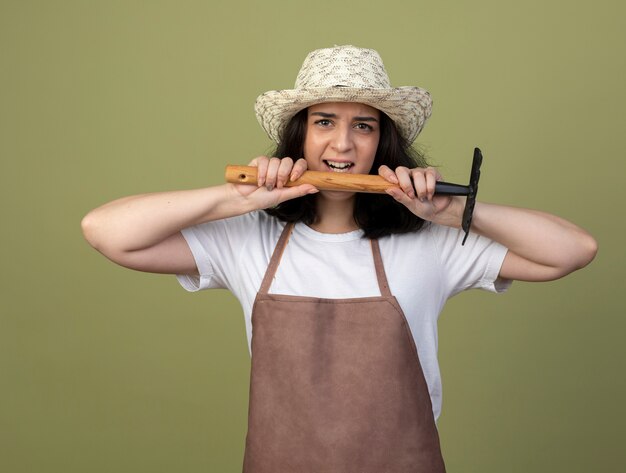 Annoyed young brunette female gardener in uniform wearing gardening hat holds rake isolated on olive green wall