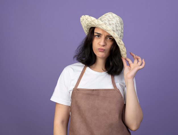 Annoyed young brunette female gardener in uniform wearing and holding gardening hat isolated on purple wall