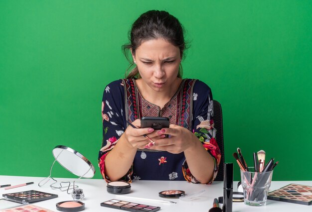 Раздраженная молодая брюнетка девушка сидит за столом с инструментами для макияжа, держа и глядя на телефон, изолированный на зеленой стене с копией пространства