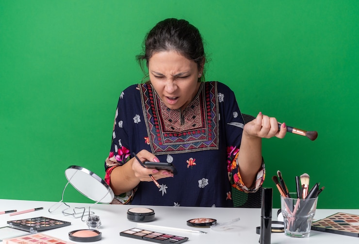 मुफ़्त फोटो annoyed young brunette girl sitting at table with makeup tools holding makeup brushes and looking at phone isolated on green wall with copy space