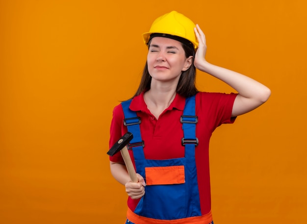 Раздраженная молодая девушка-строитель держит молоток и кладет руку на голову на изолированном оранжевом фоне с копией пространства