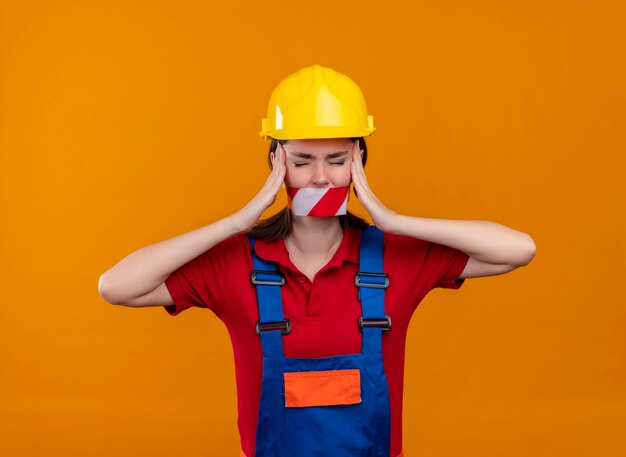 Раздраженная молодая девушка-строитель, заклеенная предупреждающей лентой, держит голову обеими руками на изолированном оранжевом фоне